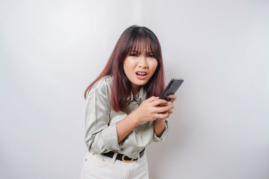A dissatisfied young Asian woman looks disgruntled wearing sage green shirt irritated face expressions holding her phone