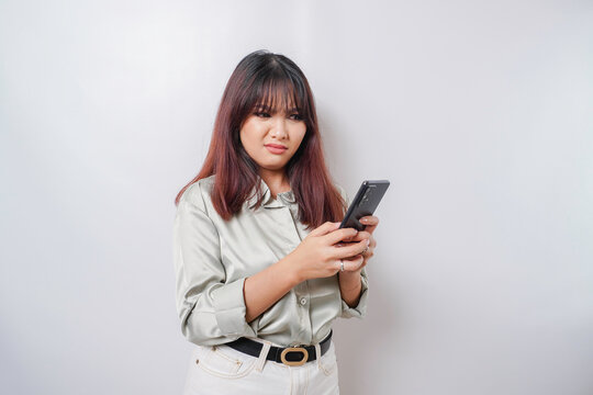 A dissatisfied young Asian woman looks disgruntled wearing sage green shirt irritated face expressions holding her phone