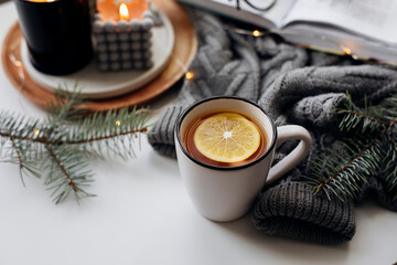Winter home cozy concept. Mug with lemon tea, open book, warm sweater, candles and fir tree....