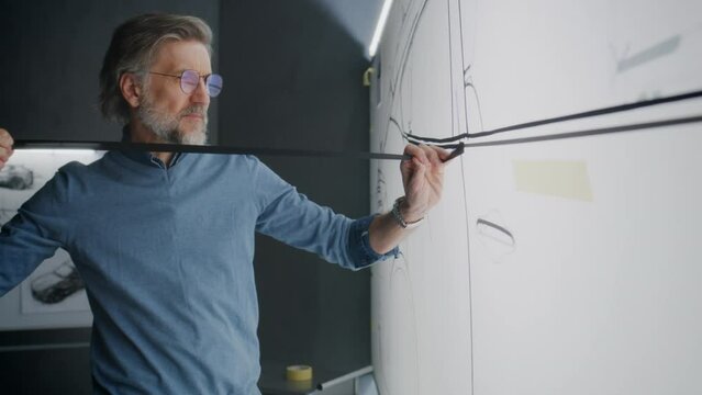 Automotive male engineer creating sketch with duct tape on paper whiteboard, developing futuristic new generation electric car. He working in modern car design development studio.