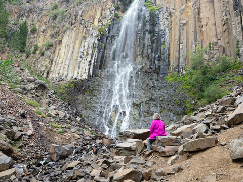 Senior Adult Woman Wearing a Fuchsia Jacket Sitting on a Boulder at the Base of a Waterfall