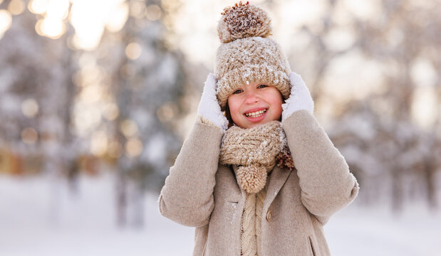 Portrait of happy cute little girl in knitted hat and warm clothes standing in snowy winter park