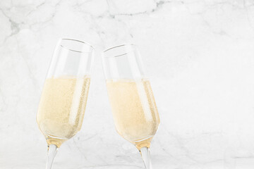 New Year festive background with bright fizz champagne in two glasses in motion of clang glasses together on soft light white marble backdrop, closeup, top part, copy space.