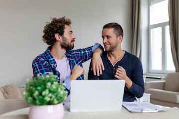 Lovely gay couple laughing together while sitting in their living room at home. Two romantic young male lovers having fun surfing the internet indoors. Young gay couple living together.