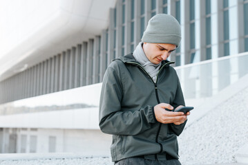 Portrait of european young man in outerwear browsing internet on smartphone, chatting while...