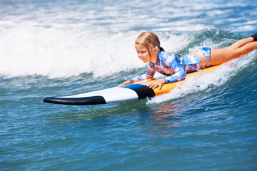 Happy baby girl - young surfer ride on surfboard with fun on sea waves. Active family lifestyle, kids outdoor water sport lessons and swimming activity in surf camp. Beach summer vacation with child.