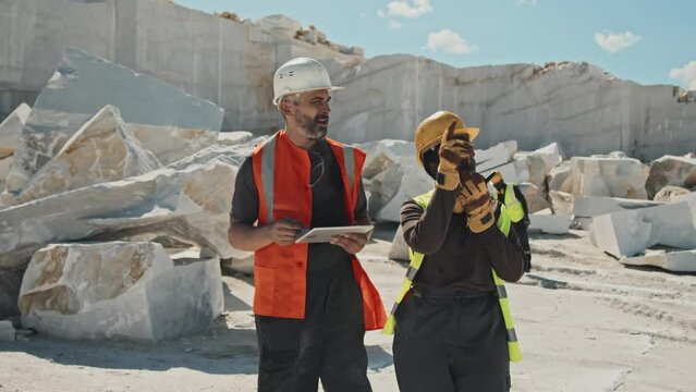 Mature Caucasian foreman and African American engineer walking along marble quarry territory discussing extraction plans