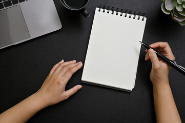 Top view, A female hand holding pen, taking notes on spiral notepad in her modern black desk