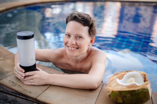A beautiful woman in the pool prepares a smoothie in a small cordless blender from coconut banana and milk. Emotional healthy smile.