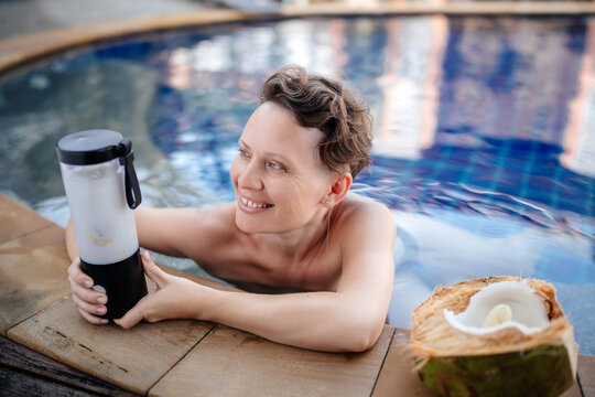 A beautiful woman in the pool prepares a smoothie while holding a small wireless blender in her hands. Healthy eating and self care.