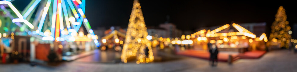 Christmas market, with blurry and defocused colorful garland lights, carousel and fir tree. Panorama