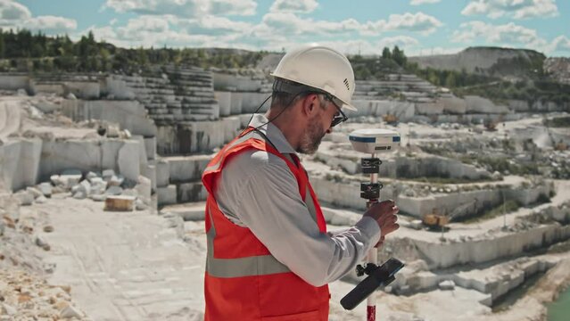 Mature Caucasian man wearing hardhat and sunglasses doing topographic survey of marble quarry using smartphone and geodetic receiver
