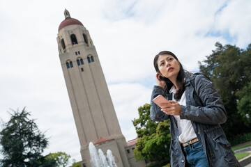 dutch angle of lost asian Japanese exchange student at school university looking into space trying to figure out how to get to classroom while consulting campus map on phone near hoover tower