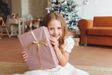 A little girl in a white dress is sitting on the floor and holding a New Year's gift against the...