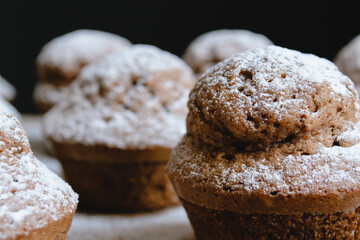 Close-up of a baked chocolate cupcake on a wooden board and sprinkled with white powdered sugar surrounded by other cupcakes