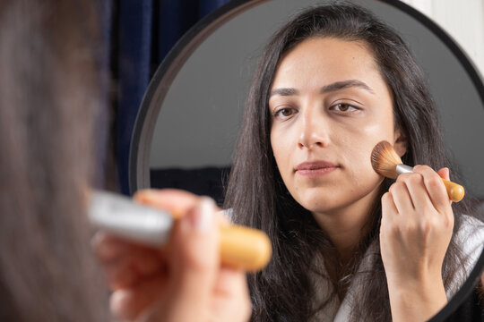 Applying liquid foundation, young beautiful woman applying liquid foundation on her face with brush. Caucasian woman looking at her reflection in the mirror. Close up photo of make up concept idea.