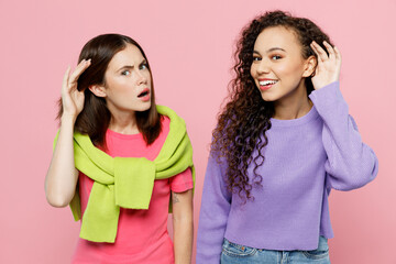 Young two friends curious nosy women wears green purple shirts looking camera together try to hear you overhear listening intently isolated on pastel plain light pink color background studio portrait.