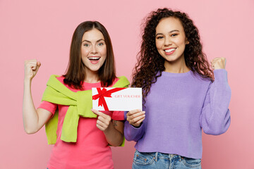 Young two friends excited happy fun women wear green purple shirts together hold gift certificate coupon voucher card for store do winner gesture isolated on pastel plain light pink color background.