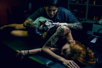 Fototapeta na wymiar Professional tattooist makes the tattoo on a girl waist, focusing on tattoo machines in a modern studio lowlight.