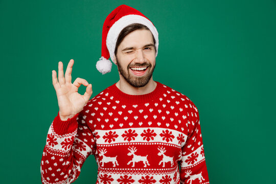 Merry Fun Young Man 20s Wear Red Warm Knitted Sweater Santa Hat Posing Showing Okay Ok Gesture Wink Isolated On Plain Dark Green Background Studio Portrait. New Year 2023 Holiday Celebration Concept.