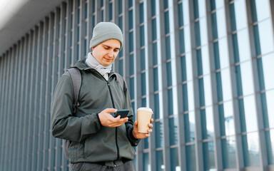 Young caucasian man in outerwear using smartphone holding cup of coffee while walking down street and looking at screen of mobile phone