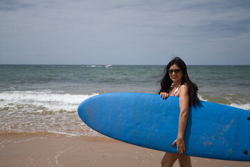 Naklejka na ściany i meble South American woman, young and beautiful, brunette with sunglasses and swimsuit, coming out of the water holding a blue surfboard. Concept sea, sand, sun, beach, vacation, surf, summer.