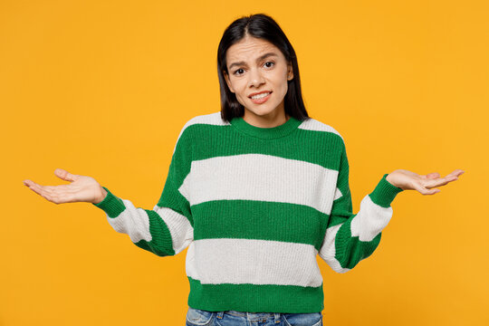 Young confused brunette latin woman wear casual cozy green knitted sweater spread hands say oops shrugging shoulders looking puzzled, have no idea isolated on plain yellow background studio portrait.