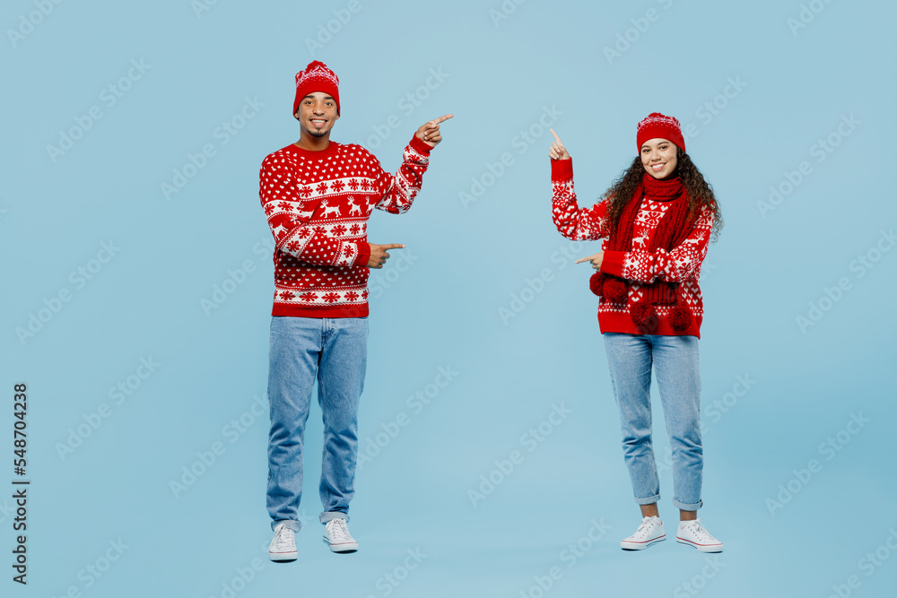 Wall mural Full body merry young couple two man woman wear red Christmas sweater Santa hat posing point finger aside on area mock up isolated on plain pastel blue background. Happy New Year 2023 holiday concept.