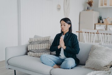Fototapeta premium Female does breathing exercises, meditating, sitting in lotus pose on couch at home. Stress relief