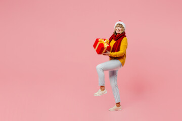 Full body elderly woman 50s year old wear yellow knitted sweater red scarf Santa hat posing hold present box gift ribbon bow isolated on plain pink background. Happy New Year Christmas 2023 concept.