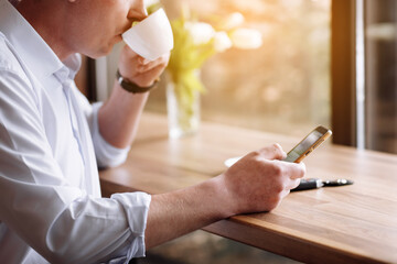 Young businessman drinking coffee and using smartphone while working in a cafe. Man with cup of coffee looking at his mobile phone during a work break in coffee shop.