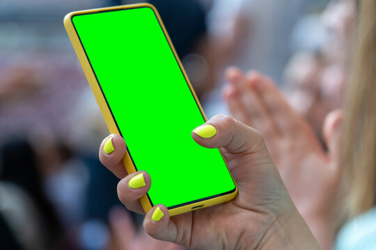Woman holds mobile smartphone with green screen in crowd at concert at stadium, sends photos to social networks at summer open-air music festival. Online internet concept communication app