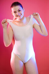 Cheerful young woman posing in white one piece swimsuit in front of pink background in studio	
