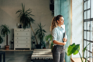 Adult businesswoman standing at the office, looking through the window, enjoying the view.