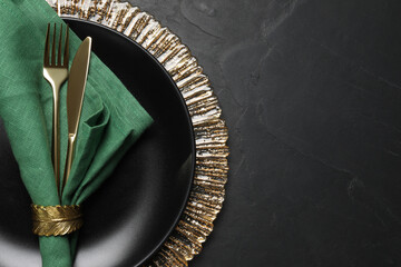Plate with green fabric napkin, decorative ring and cutlery on black table, top view. Space for text