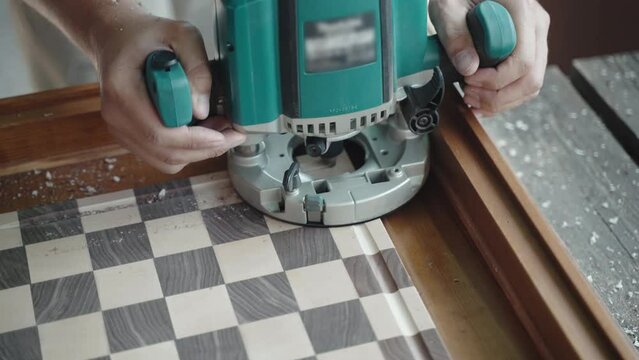 Professional carpenter using grinding machine for carving holes on the sides of wooden board, joiner assistant removing sawdust with vacuum cleaner. Process of making final design of two colour wooden