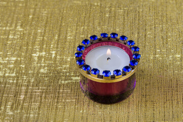 A tea light with burning flame in a blue purple glass holder on a golden textured background.