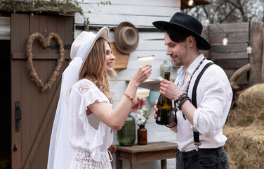 country bride in a white long dress and hat and groom in a white shirt drinking champagne in the village wedding, boho style.
