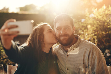 Happy couple, selfie on a phone with a kiss for love, care and support while outdoor in garden with lens flare for summer. Man and woman together with smartphone for social media profile picture