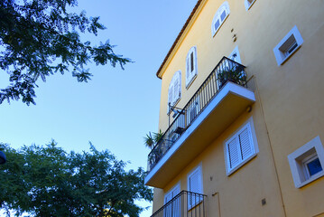 Balcony with windows on a residential building. Yellow building facade against trees with green leaves. Buildings architecture and house facade. Modern minimalistic architecture in Spain.