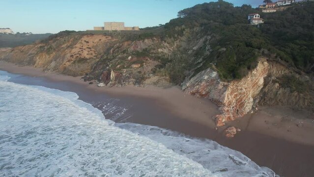 Aerial view from drone in Bidart France of a man surfer comes to get his surfboard in the hollow of the coastal cliff when the tide is rising and joins the abandoned building on the beach at sunset