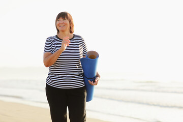 Middle age woman holding a sports mat and preparing to practice yoga outdoors on sea beach. Happy mature overweight woman exercising on seashore. copy space. Meditation, yoga and relaxation concept.