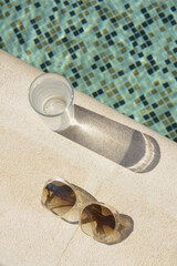 Stylish sunglasses and glass of water near outdoor swimming pool on sunny day, flat lay