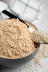 Sesame flour in ceramic bowl and spoon of seeds on grey table, closeup