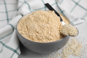 Sesame flour in ceramic bowl with spoon of seeds on grey table, closeup
