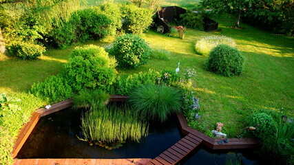 attraktiver naturbelassener Garten mit Brücke an roter Terrasse angrenzender Teich im Frühling