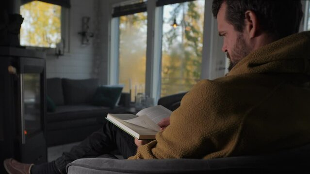 Caucasian man reading a book in a warm cottage in the woods. Static view