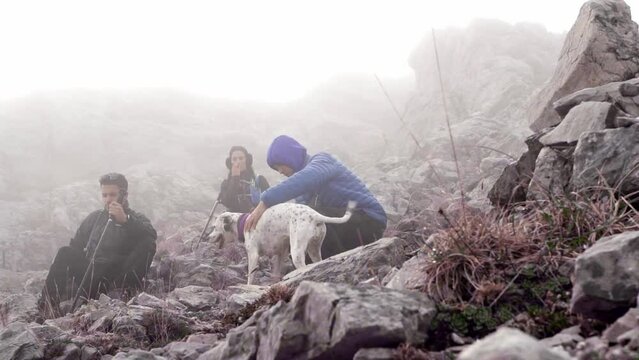 The three young hikers and their two dogs rest while climbing a rocky mountain, using their poles to help each other and equipped with mountain clothing. The day is cloudy and cold.