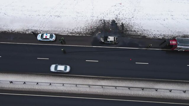 Losses - a burned-out car on the edge of the motorway, aerial view. Near the fire and police cars. Help and rescue concept, 911 service. White snow on the side of the road. UHD 4K