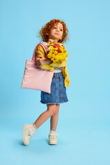 Portrait of cute stylish little girl, child with curly red hair posing with flowers isolated over blue background